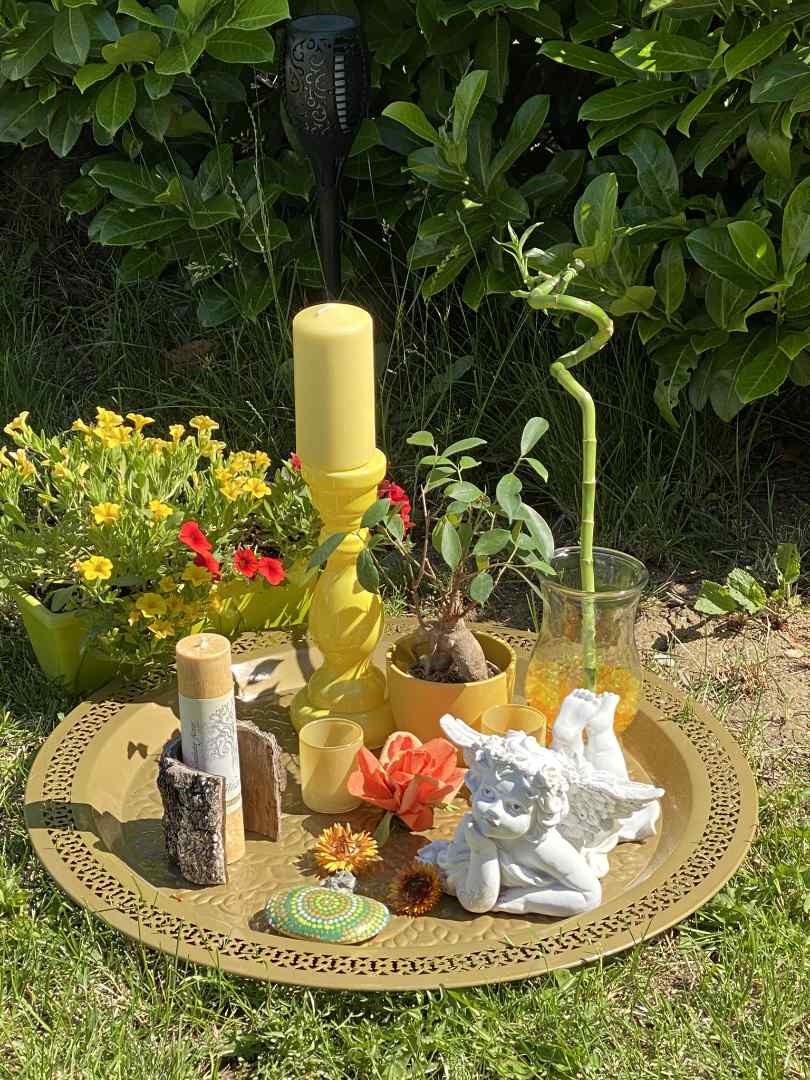 Sommer-Rauhnächte_web Dekoratives Arrangement im Garten mit gelben Kerzen, Blumen, einem weißen Engelsfigur und symbolischen Naturmaterialien auf einem goldenen Tablett – gestaltet für die Sommerrauhnächte.